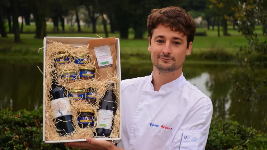 Coffret gourmand ouvert, bocaux et bouteilles calés dans la paille, présenté par un membre de l'équipe Panier de Région